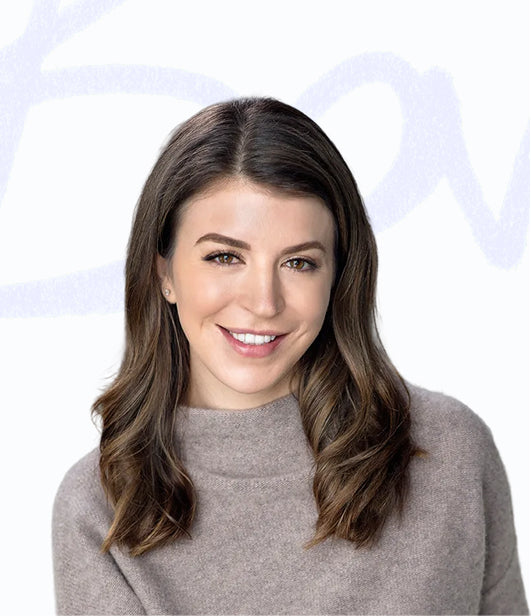 Woman with wavy brown hair smiling in gray sweater against light background
