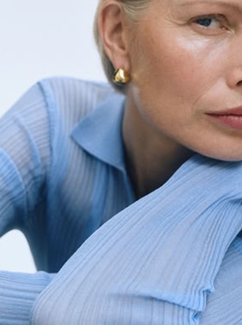 Woman wearing gold stud earrings and light blue ribbed blouse, elegant minimalist style